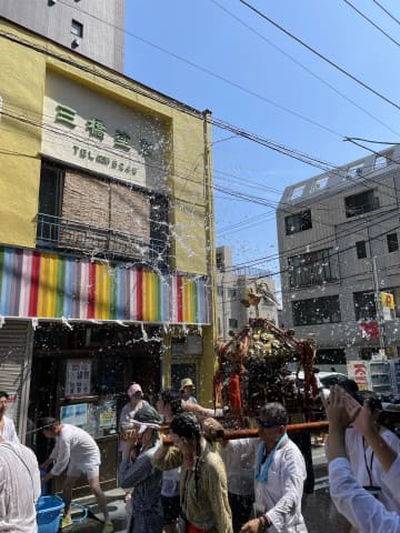 宮川町の黄色い社屋の前を神輿が通り、担ぎ手たちに水しぶきが上がる夏祭りの様子