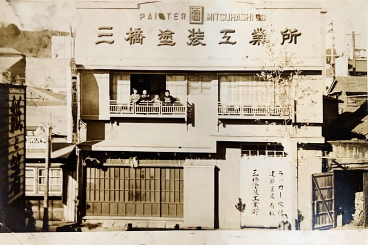 昭和初期の三橋塗装工業所の社屋。正面に大きな社名看板を掲げ、二階の手すりから家族が顔を出している白黒写真
